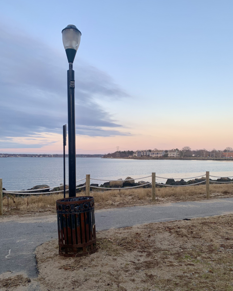 There is an ocean cove in the background. In the foreground is a light post and trash can next to a paved walking path.
